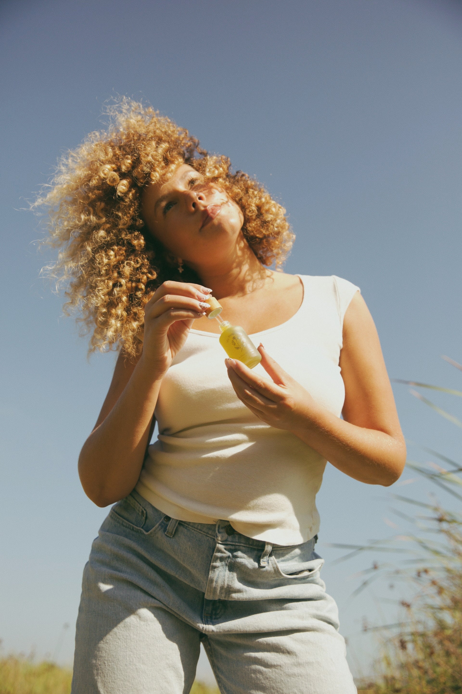 A person with curly hair, in a white sleeveless top and light jeans, stands outdoors under a blue sky, holding My Neighbor’s Tallow Blue Moon Face Serum—ideal for sensitive skin. Tall grass gently sways behind them.