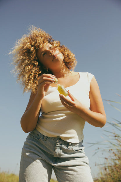 A person with curly hair, in a white sleeveless top and light jeans, stands outdoors under a blue sky, holding My Neighbor’s Tallow Blue Moon Face Serum—ideal for sensitive skin. Tall grass gently sways behind them.
