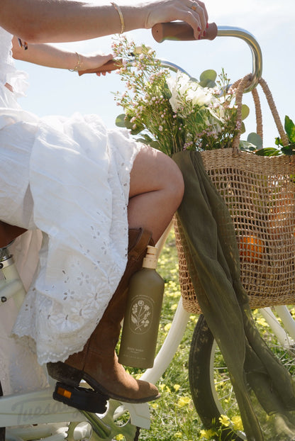 A woman in a white dress and brown boots rides a white bike with flowers and My Neighbor’s Tallow Wild Bergamot Body Wash in the basket, enjoying a sunny day in a grassy field.