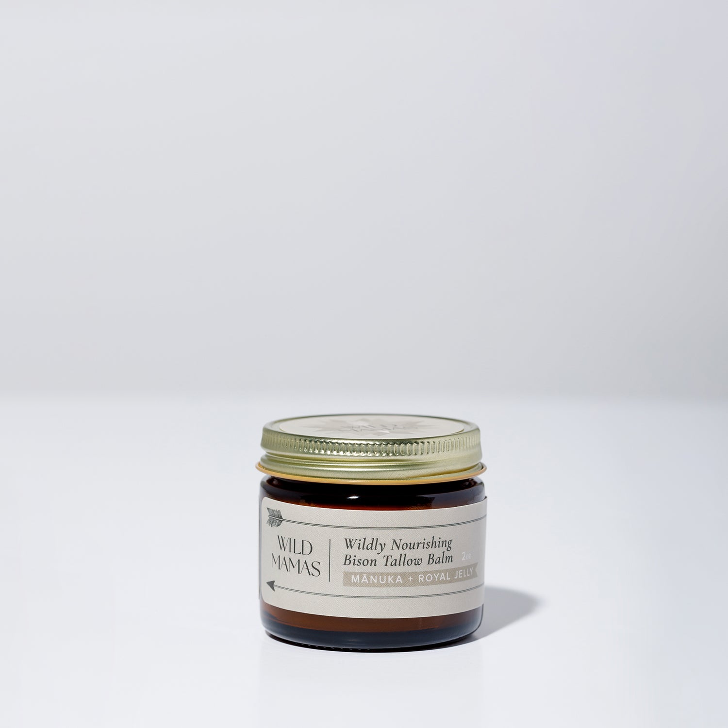A small glass jar with a gold lid, labeled "Wildly Nourishing Bison Tallow Balm - Mānuka Honey + Royal Jelly" by WildMamas, sits on a plain white background. This balm delivers nutrient-rich bison tallow for regenerative skincare.