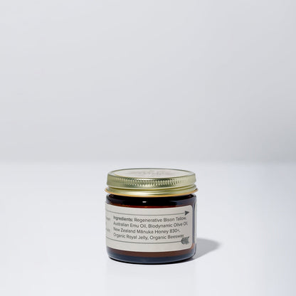 A small brown glass jar with a gold lid from WildMamas sits on a white surface. The label reads: Wildly Nourishing Bison Tallow Balm – Mānuka Honey + Royal Jelly, made with bison tallow, emu oil, olive oil, beeswax, and more.
