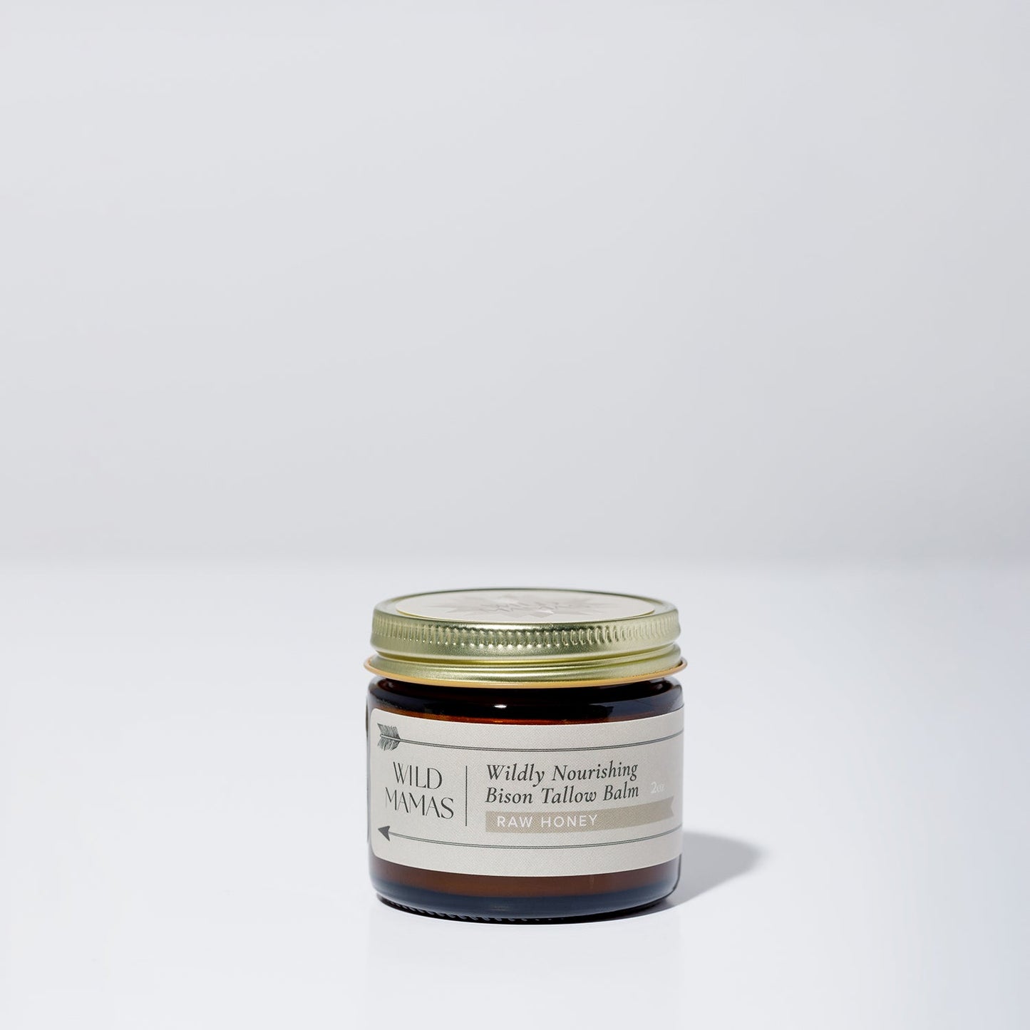 A small glass jar with a gold lid contains Wildly Nourishing Bison Tallow Balm - Raw Honey by WildMamas, made from regenerative sources. The label shows the Raw Honey scent, and the jar rests on a simple, light background.