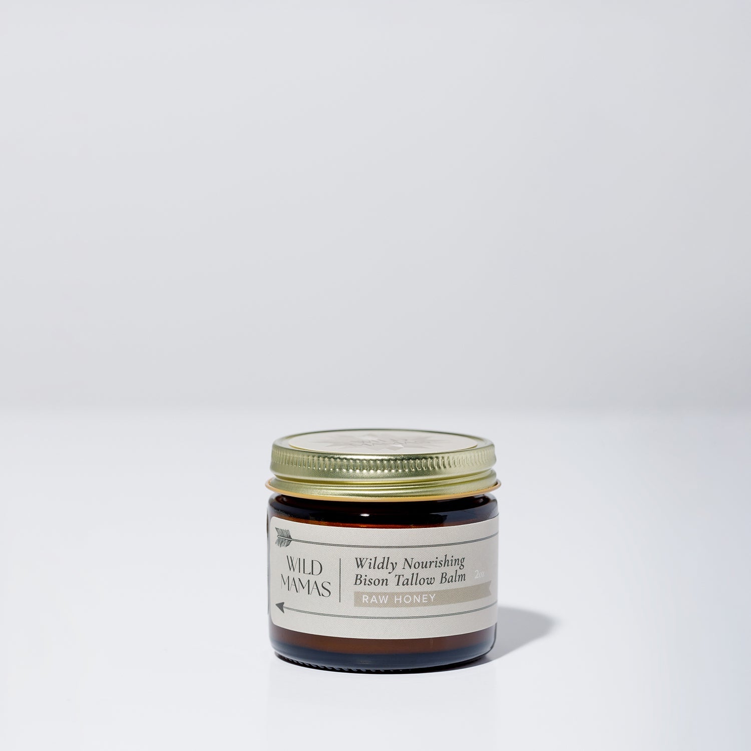 A small glass jar with a gold lid contains Wildly Nourishing Bison Tallow Balm - Raw Honey by WildMamas, made from regenerative sources. The label shows the Raw Honey scent, and the jar rests on a simple, light background.