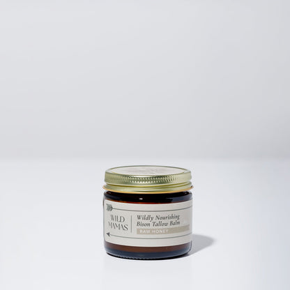 A small glass jar with a gold lid contains Wildly Nourishing Bison Tallow Balm - Raw Honey by WildMamas, made from regenerative sources. The label shows the Raw Honey scent, and the jar rests on a simple, light background.