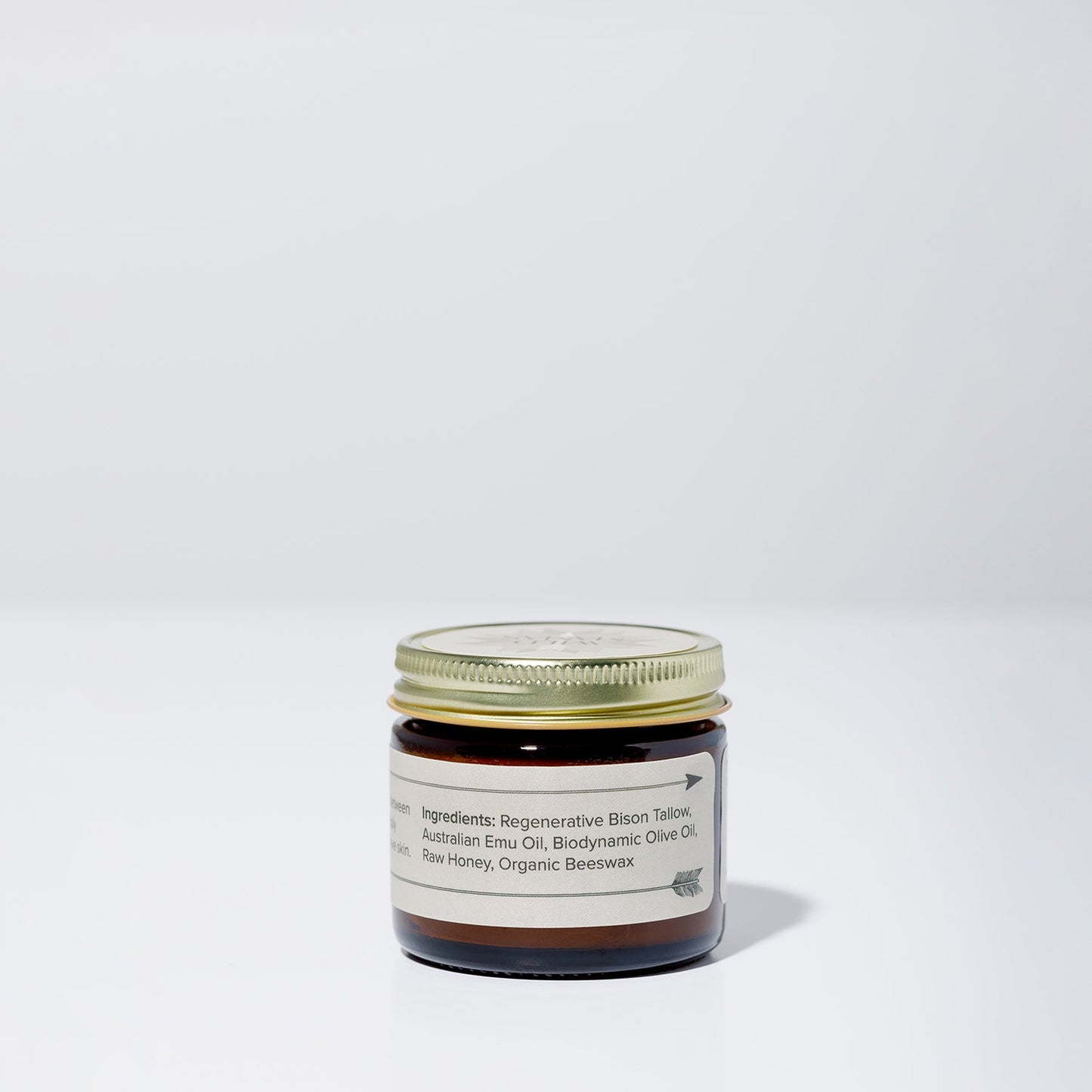 A small brown glass jar with a gold lid sits on a white surface. The label reads “WildMamas Wildly Nourishing Bison Tallow Balm - Raw Honey,” featuring bison tallow, emu oil, olive oil, honey, and beeswax. The background is plain light gray.