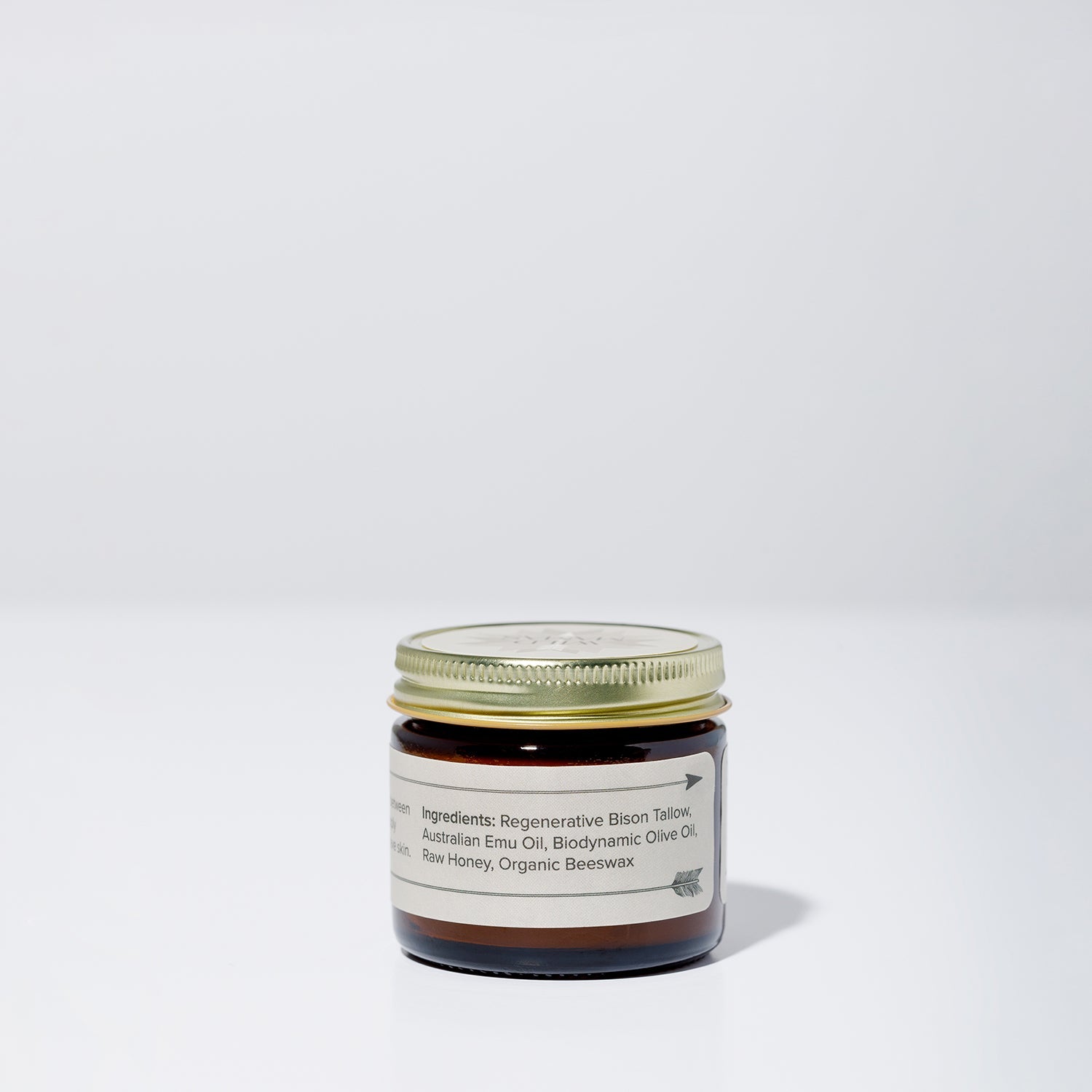 A small brown glass jar with a gold lid sits on a white surface. The label reads “WildMamas Wildly Nourishing Bison Tallow Balm - Raw Honey,” featuring bison tallow, emu oil, olive oil, honey, and beeswax. The background is plain light gray.