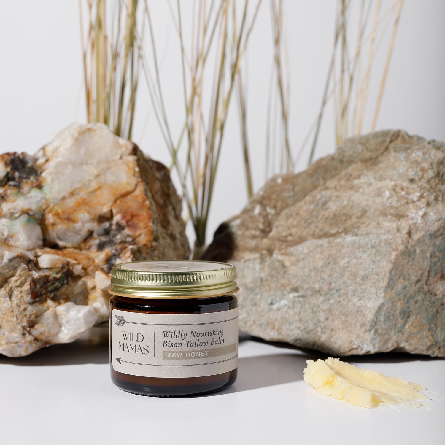 A glass jar of WildMamas Wildly Nourishing Bison Tallow Balm - Raw Honey rests on a white surface next to a dollop of balm, showcasing regenerative bison tallow skincare with rocks and dried grass in the background.