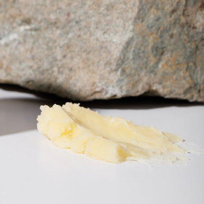 A smear of pale yellow Wildly Nourishing Bison Tallow Balm - Raw Honey by Wild Mamas rests on a white surface before a rough gray stone, highlighting the richness of regenerative bison tallow skincare.