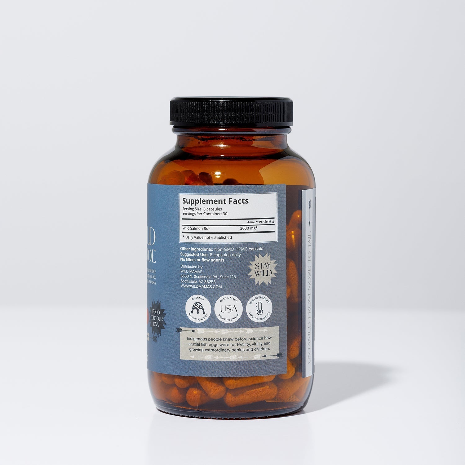 A brown glass bottle with a black lid contains orange WildMamas Wild Salmon Roe capsules. The label shows supplement facts, certifications, and icons for vegan, made in the USA, and "Stay Wild." The bottle is on a plain white background.