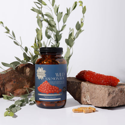 A jar of WildMamas Wild Salmon Roe sits on a white surface with greenery and rocks, while salmon roe and Omega-3-rich yellow capsules rest nearby, showcasing this superfood in a clean, minimalistic setting.