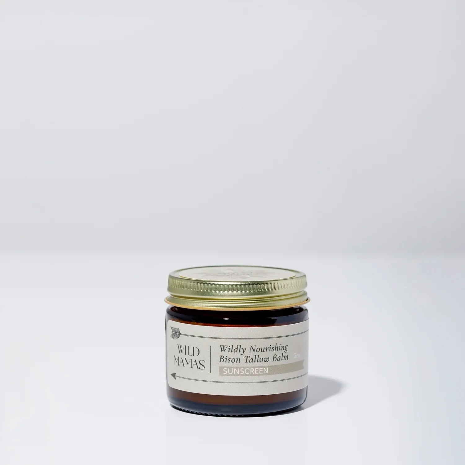 A small brown glass jar with a gold lid, labeled "Wildly Nourishing Bison Tallow Balm - Sunscreen" by Wild Mamas, sits on a plain white background—a natural choice for regenerative skincare.