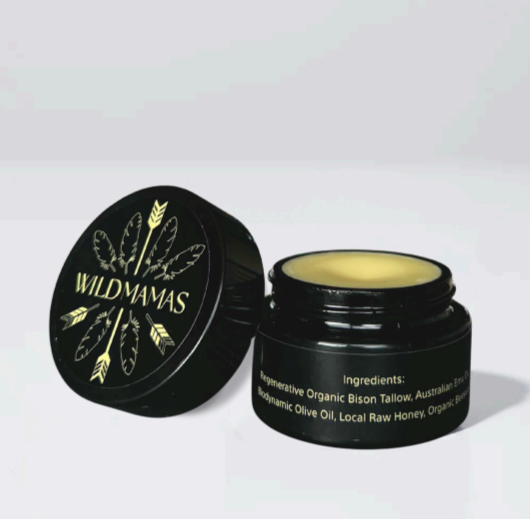 An open black jar labeled "Wild Mamas" with a gold feather reveals creamy Wildly Nourishing Bison Tallow Balm - Raw Honey, made with bison tallow, emu oil, olive oil, honey, and beeswax for nourishing skincare.