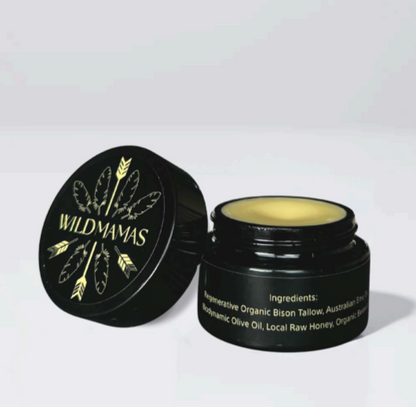 An open black jar labeled "Wild Mamas" with a gold feather reveals creamy Wildly Nourishing Bison Tallow Balm - Raw Honey, made with bison tallow, emu oil, olive oil, honey, and beeswax for nourishing skincare.