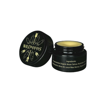 An open small black jar of Wild Mamas' Wildly Nourishing Bison Tallow Balm – Raw Honey, with an illustrated lid of leaves and feathers. Yellow balm is visible inside, and ingredients appear in white text on the side.