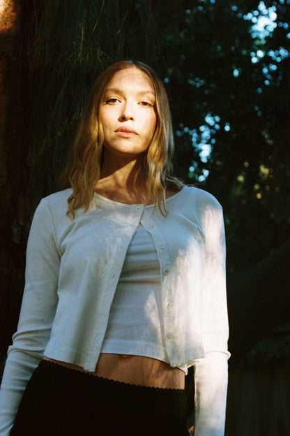 Ivory 
A woman with long, wavy hair stands in a forest wearing an LA Relaxed Organic Rib Cardigan over a white top. Sunlight filters through the trees, creating patterns on her as she gazes calmly at the camera, embodying a serene 90s closet vibe.