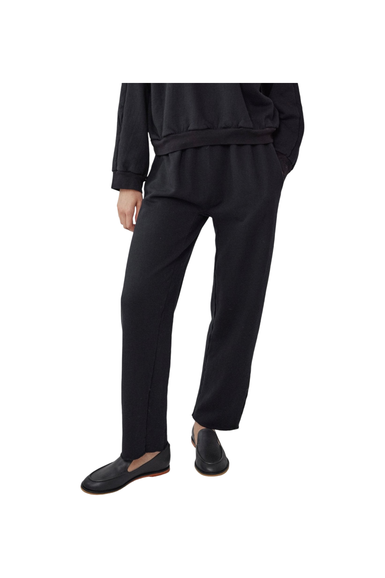 An individual is dressed in a chic black ensemble that includes the Easy Sweatpant Black, an organic cotton fleece piece with an elastic waist by Wol Hide, complemented by black slip-on shoes against a black backdrop. Made in USA.