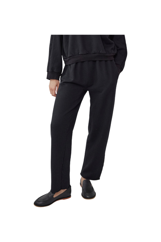 An individual is dressed in a chic black ensemble that includes the Easy Sweatpant Black, an organic cotton fleece piece with an elastic waist by Wol Hide, complemented by black slip-on shoes against a black backdrop. Made in USA.