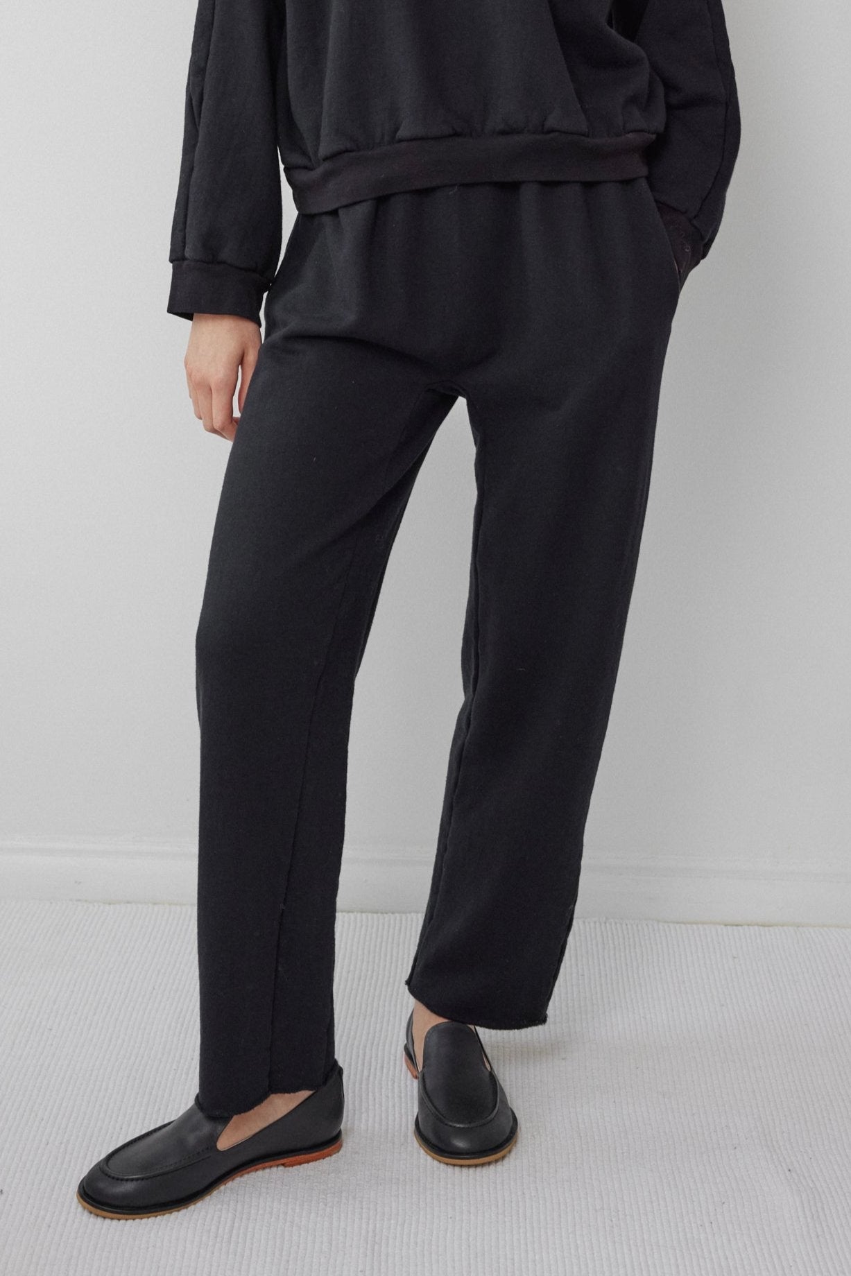 A person dressed in the Wol Hide Easy Sweatpant Black, paired with a black sweatshirt made from USA-made organic cotton fleece, stands against a white wall. They are also wearing black loafers, with their left hand tucked into their pocket while the right hand remains out of sight.