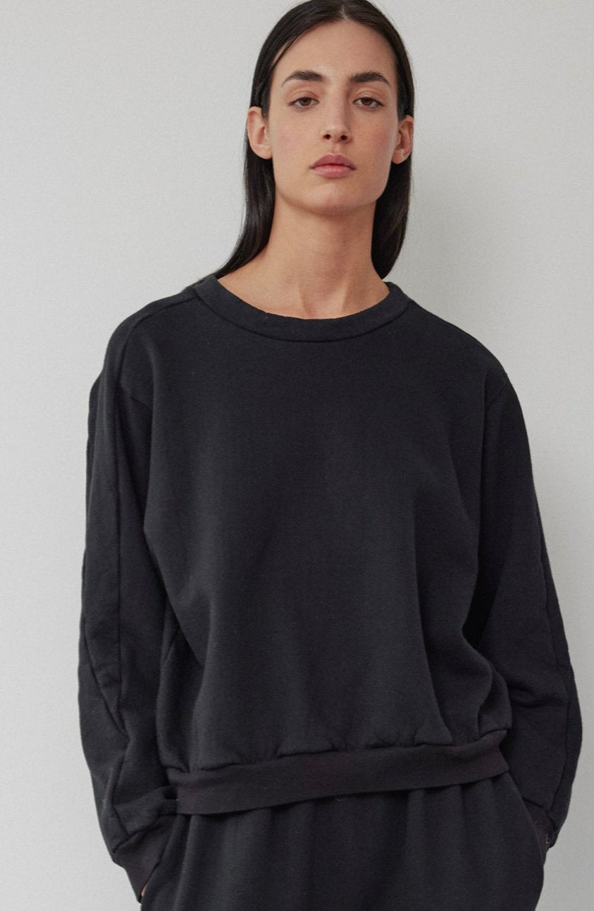 A person with long dark hair stands against a plain light background, wearing an Easy Winter Sweatshirt in black from Wol Hide, accompanied by pants. The loose-fitting boxy crew neck sweatshirt is made in the USA and crafted from organic cotton fleece, exuding a relaxed vibe. Their expression is neutral, with hands tucked into their pockets.