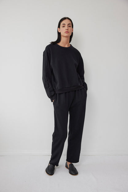 A person with long, dark hair is standing in a relaxed pose against a plain white wall. They are wearing a black sweatshirt made from organic cotton fleece, paired with Wol Hide's Easy Sweatpant Black featuring an elastic waist, and slip-on shoes. Their hands are in their pockets, exuding a casual, comfortable vibe.