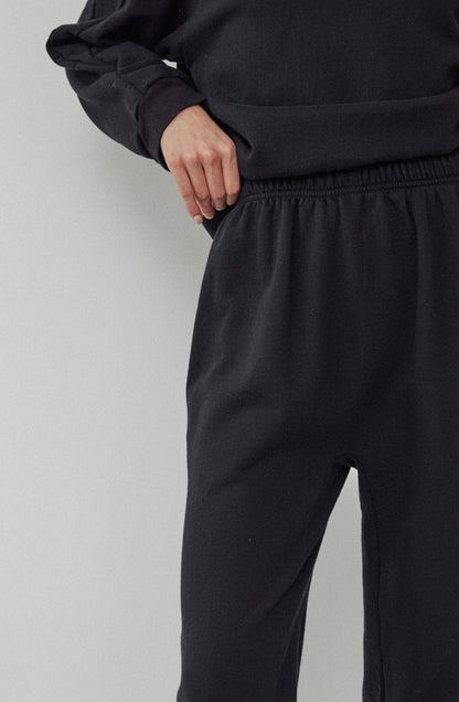 A person is wearing Wol Hide's Easy Sweatpant Black, paired with a loose black sweatshirt, standing against a light gray wall. Crafted from organic cotton fleece, this USA-made outfit highlights comfort and style with its sustainable design. Their left hand rests on their hip, emphasizing the ensemble.
