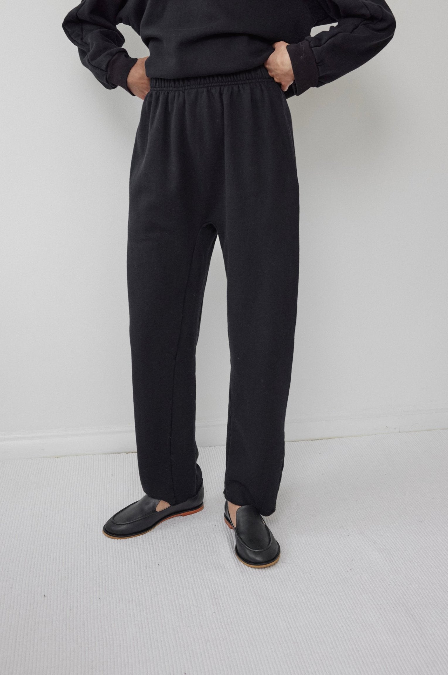 A person stands against a white background, wearing a black organic cotton fleece long-sleeve shirt and matching Wol Hide's Easy Sweatpant in black with an elastic waist. Their hands are in their pockets, complemented by black slip-on shoes.