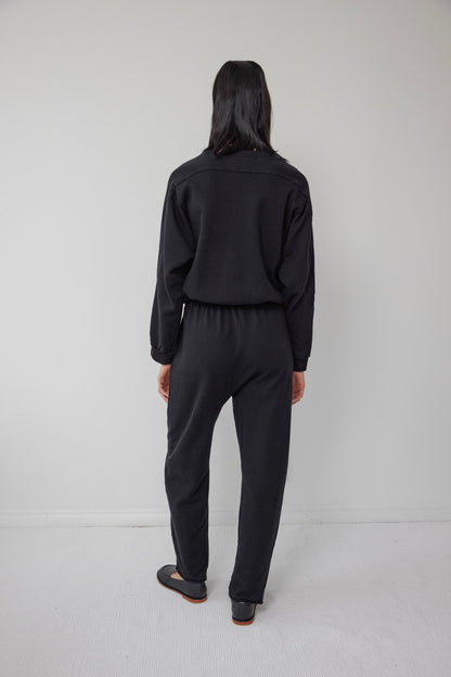 A person with long dark hair stands facing a light-colored wall. They are dressed in a USA-made, black jumpsuit featuring an elastic waist, complemented by Wol Hide's Easy Sweatpant Black and matching black shoes. The floor is white.