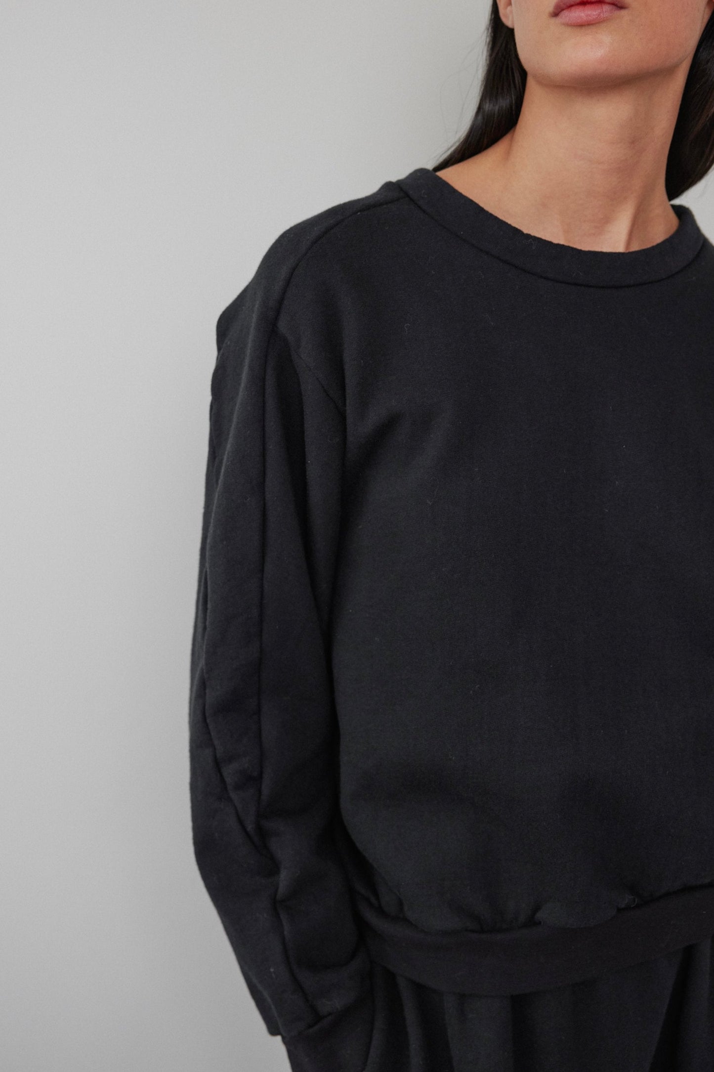 A person wearing the Wol Hide Easy Winter Sweatshirt in Black, crafted from organic cotton fleece, stands against a light gray background. The image highlights the sweatshirt's simple, casual design and upper body, with the person's face partially out of frame.