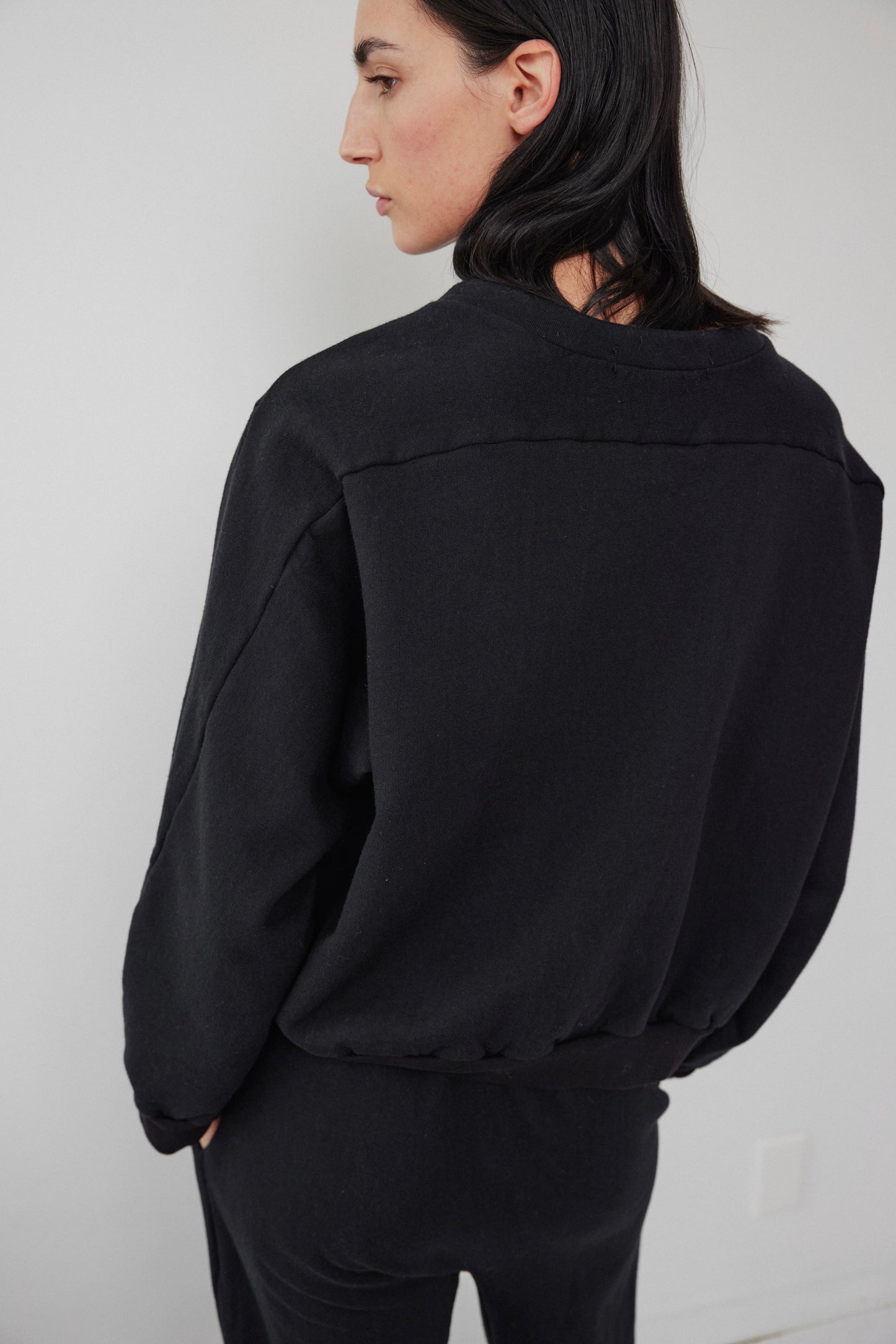 A person with long dark hair is standing in a relaxed pose, facing away from the camera. They are wearing the Wol Hide Easy Winter Sweatshirt in black and black pants, set against a plain white background.