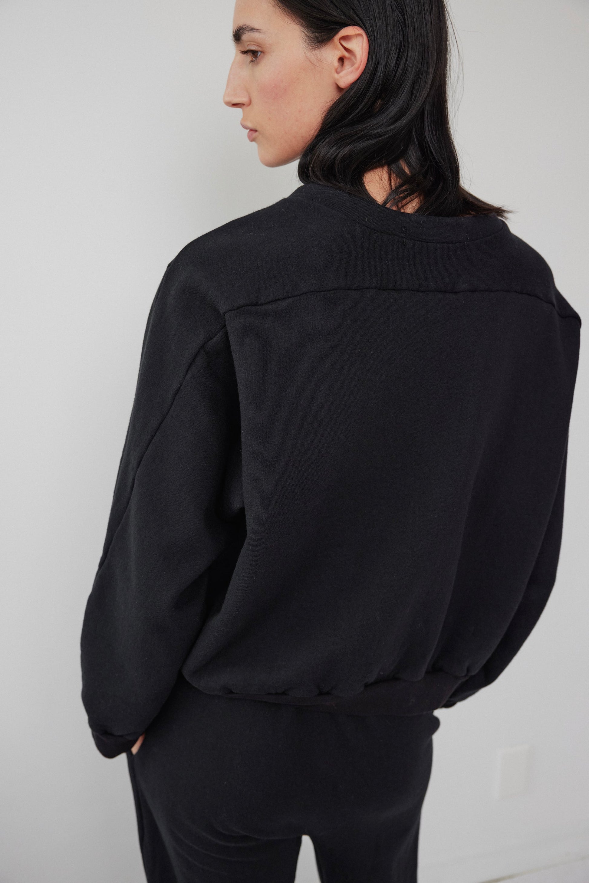 A person with long dark hair is standing in a relaxed pose, facing away from the camera. They are wearing the Wol Hide Easy Winter Sweatshirt in black and black pants, set against a plain white background.