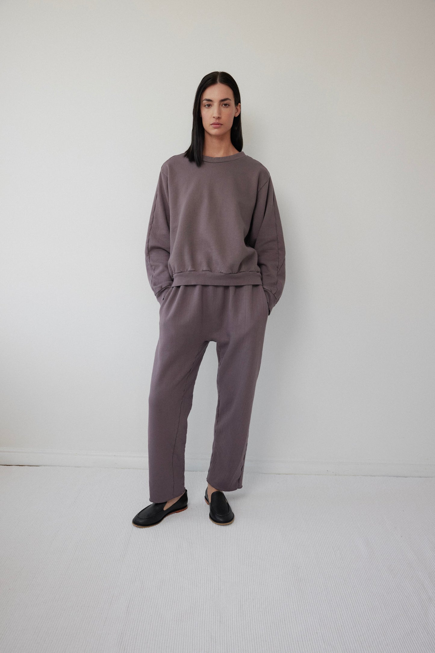 A person with long dark hair stands against a white wall, wearing Wol Hide's Easy Winter Sweatshirt in Mushroom and loose-fitting pants made of organic cotton fleece. Hands in pockets, they complete the relaxed yet stylish look with black slip-on shoes.