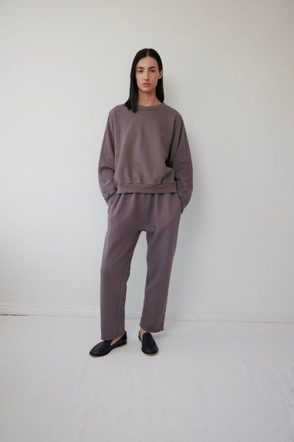 A person with long dark hair stands against a white wall, wearing Wol Hide's Easy Winter Sweatshirt in Mushroom and loose-fitting pants made of organic cotton fleece. Hands in pockets, they complete the relaxed yet stylish look with black slip-on shoes.