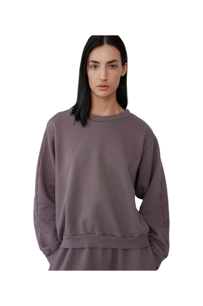 A person with long, dark hair is wearing the Wol Hide Easy Winter Sweatshirt in Mushroom, made in the USA. Their hands are tucked into matching pants crafted from 100% organic cotton. The background is plain and black.