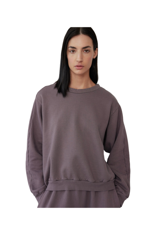A person with long, dark hair is wearing the Wol Hide Easy Winter Sweatshirt in Mushroom, made in the USA. Their hands are tucked into matching pants crafted from 100% organic cotton. The background is plain and black.