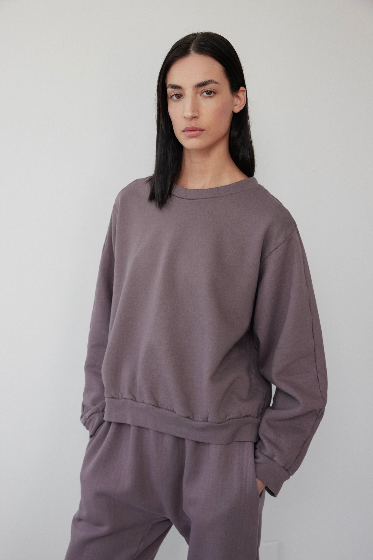 A person with long black hair wears a Wol Hide Easy Winter Sweatshirt in Mushroom and matching pants. Made in the USA, this organic cotton fleece ensemble complements their neutral expression as they stand against a plain, light-colored background with hands in pockets.