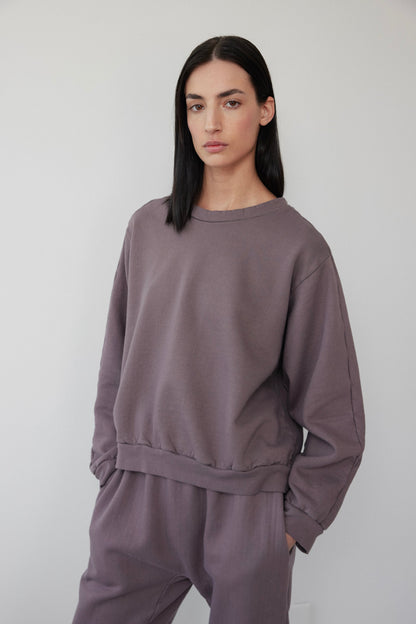 A person with long black hair wears a Wol Hide Easy Winter Sweatshirt in Mushroom and matching pants. Made in the USA, this organic cotton fleece ensemble complements their neutral expression as they stand against a plain, light-colored background with hands in pockets.