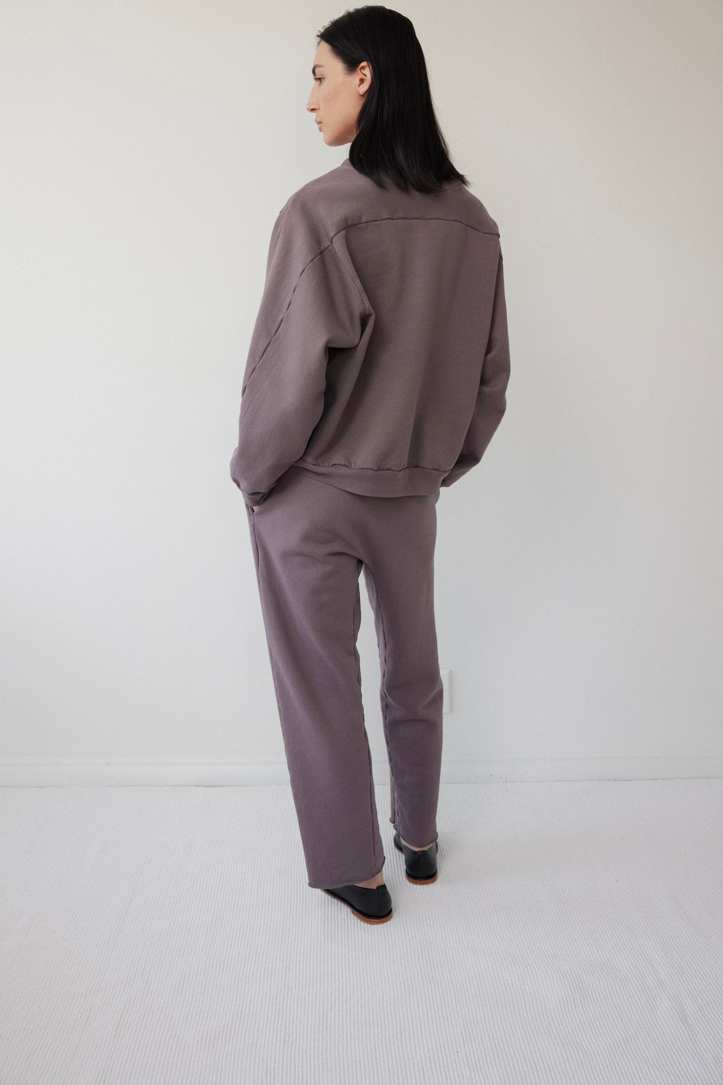In a minimalist room with light-colored walls and flooring, a person wearing Wol Hide's Easy Winter Sweatshirt in Mushroom stands gazing left. The loose-fitting muted purple crew neck made of organic cotton fleece perfectly complements their straight black hair turned away.