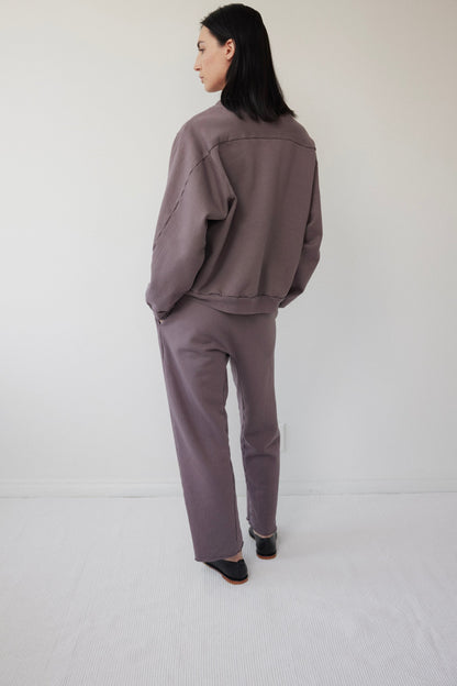 In a minimalist room with light-colored walls and flooring, a person wearing Wol Hide's Easy Winter Sweatshirt in Mushroom stands gazing left. The loose-fitting muted purple crew neck made of organic cotton fleece perfectly complements their straight black hair turned away.