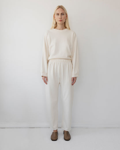 Against a white background, a person with long blonde hair models Wol Hide’s cream-colored long-sleeve shirt and Easy Sweatpant in Natural, both made from organic cotton fleece. The pants feature an elastic waist and are paired perfectly with brown shoes to complete the look.