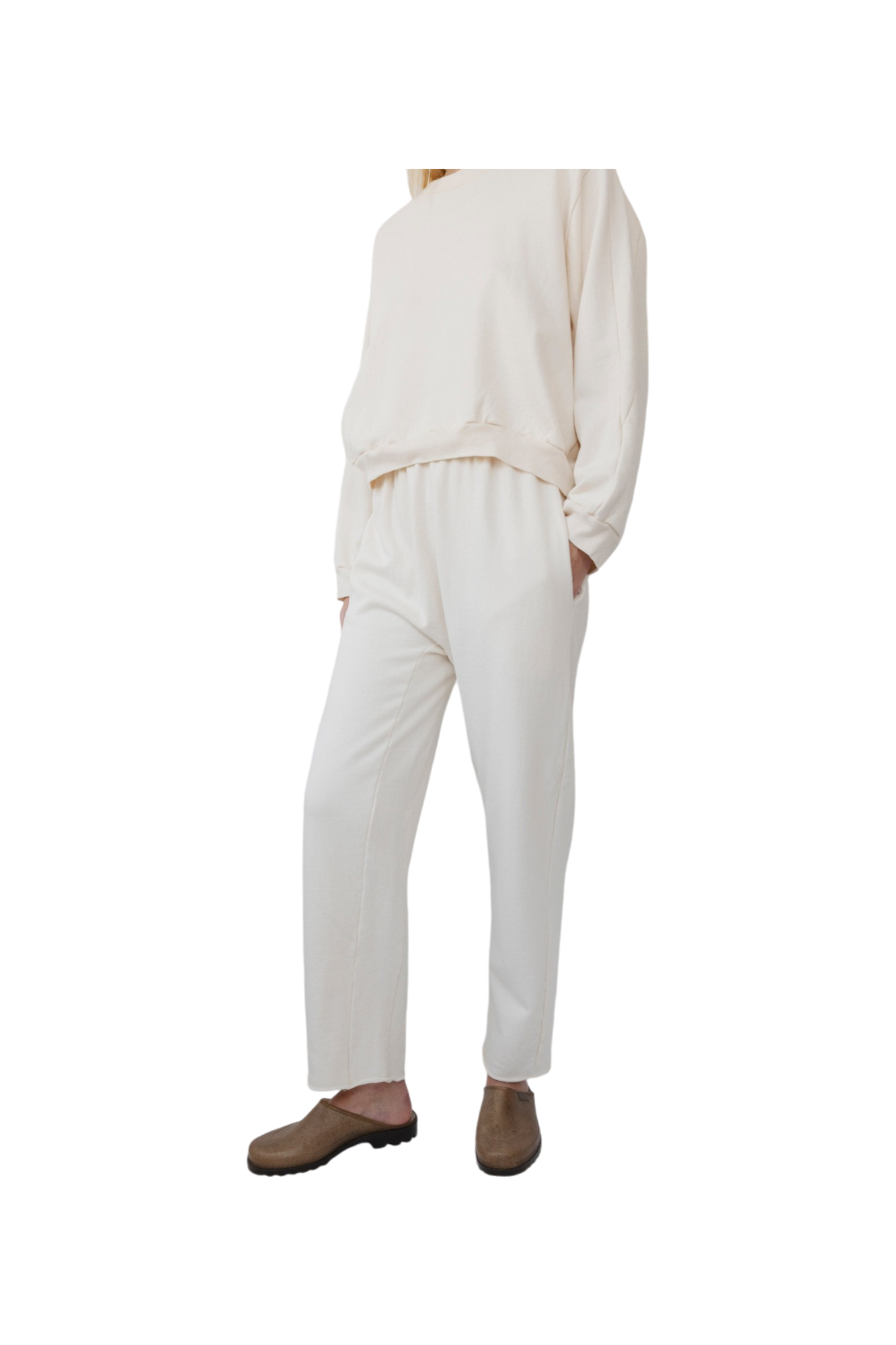 A person is wearing the Easy Sweatpant in Natural by Wol Hide, made from organic cotton fleece, consisting of a loose-fitting top and elastic waist pants, with hands in their pockets. They are also wearing brown slip-on shoes against a plain white background.