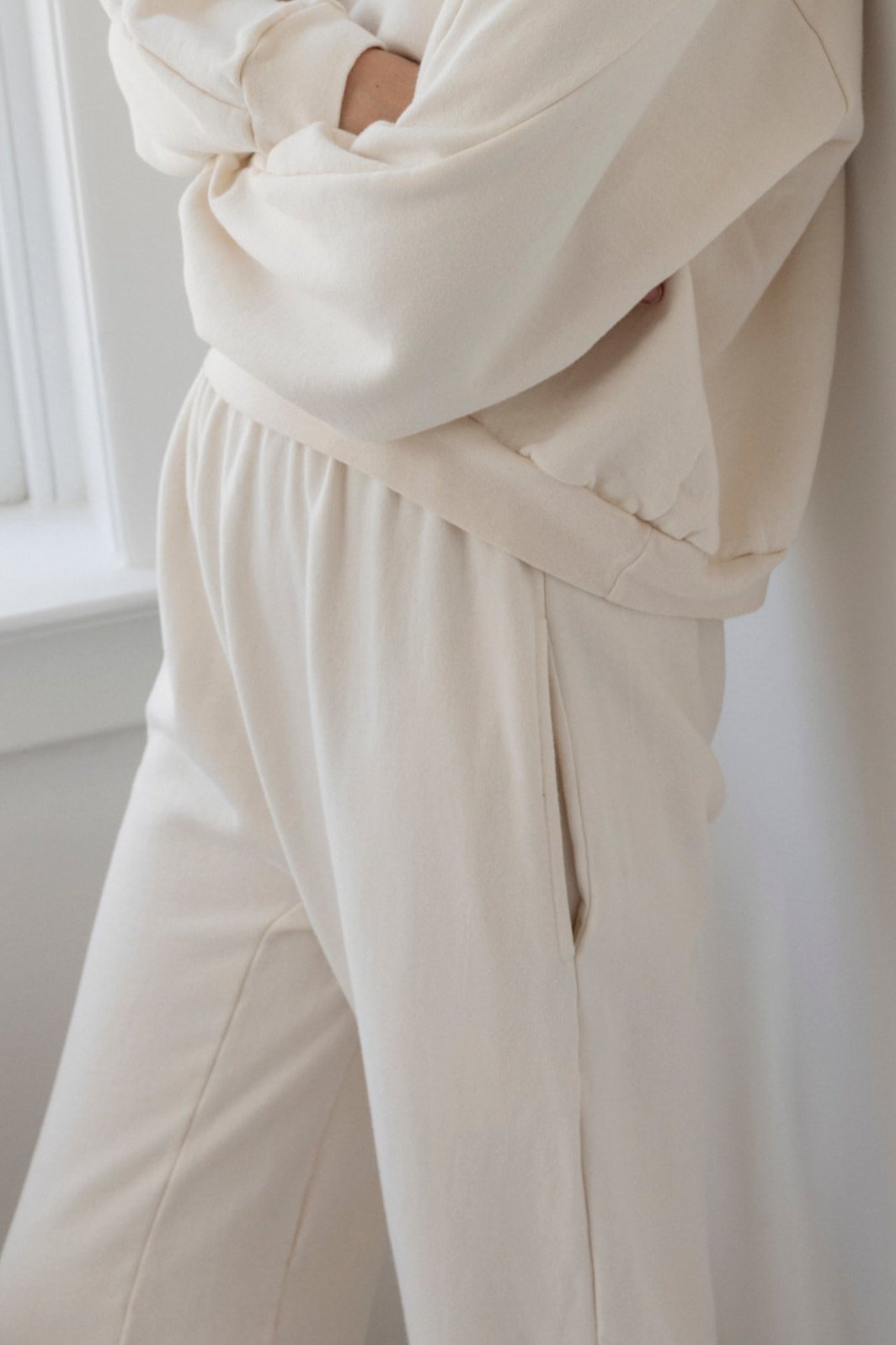 A person wearing the Wol Hide Easy Sweatpant in Natural, an oversized light beige sweatshirt, both crafted from 100% organic cotton fleece, is standing with arms crossed in front of a window.