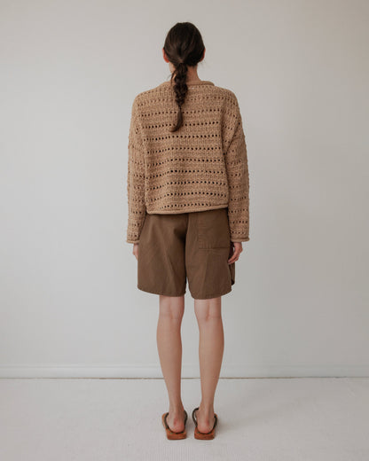 A woman with a braid stands against a white wall, facing away. She wears the Wol Hide Open Pullover in Tannin, an artisan-crafted, brown crocheted sweater made from organic cotton, paired with brown shorts and sandals.