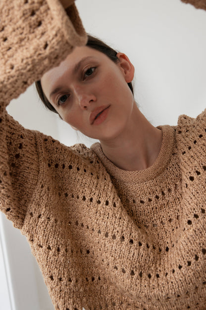 A person wearing a Wol Hide Open Pullover in Tannin, crafted from organic cotton with a delicate pattern, gazes directly at the camera. The low-angle shot captures their introspective, relaxed expression against a softly lit, plain backdrop.