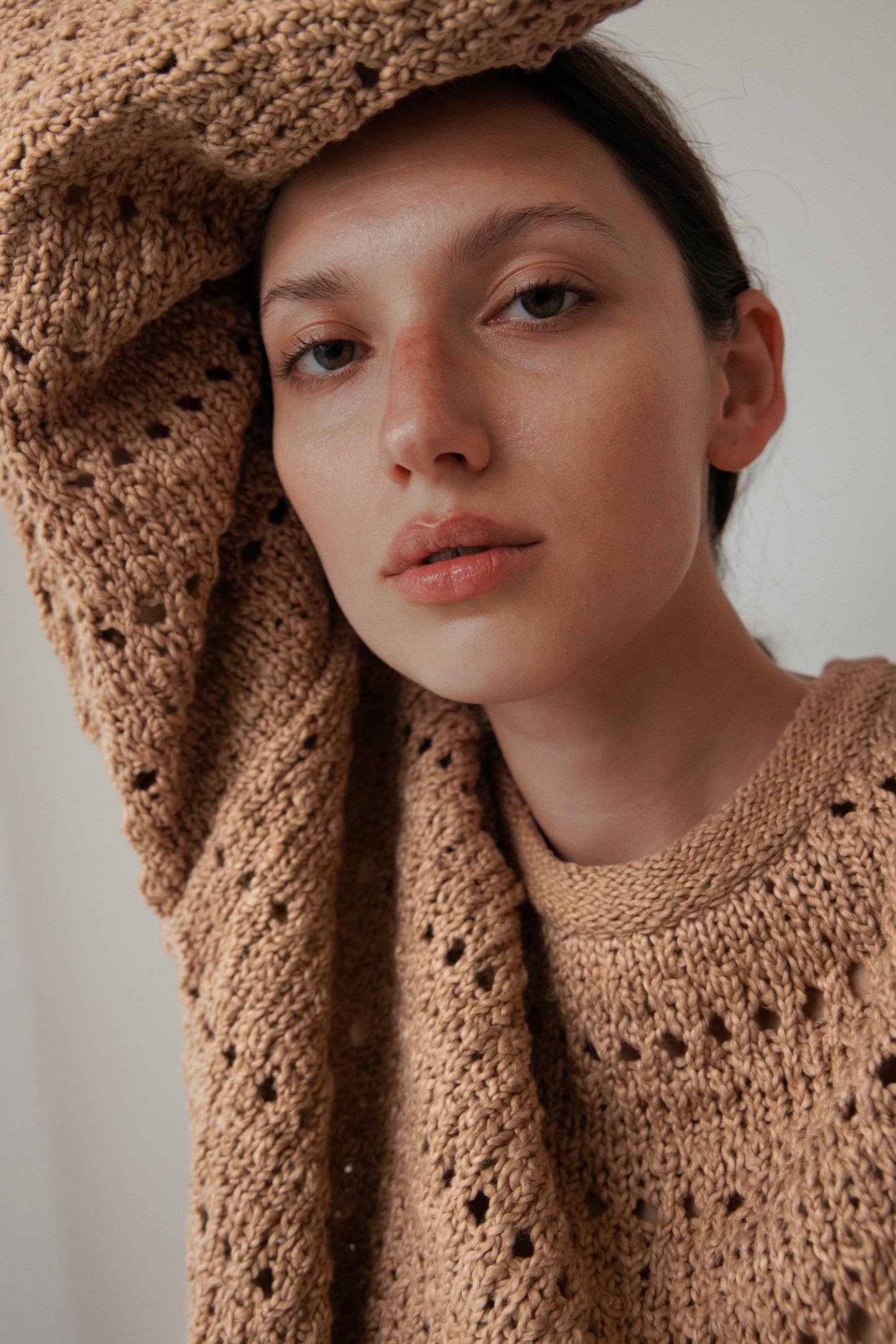 The woman, donning a Wol Hide textured Open Pullover in Tannin, crafted from plant-dyed organic cotton, leans against the wall with one arm elegantly raised overhead. Her expression is relaxed against a soothing neutral background.