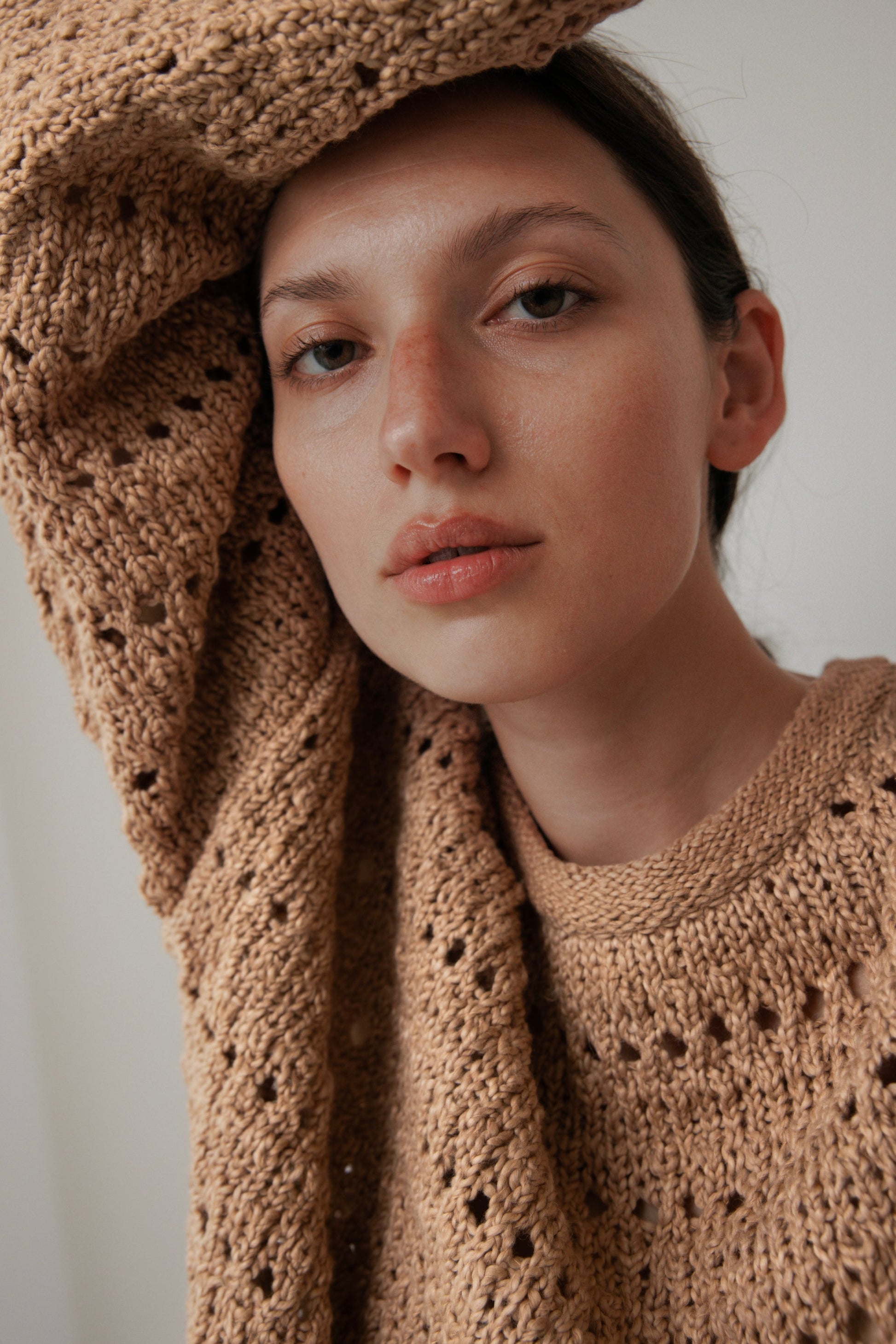 The woman, donning a Wol Hide textured Open Pullover in Tannin, crafted from plant-dyed organic cotton, leans against the wall with one arm elegantly raised overhead. Her expression is relaxed against a soothing neutral background.
