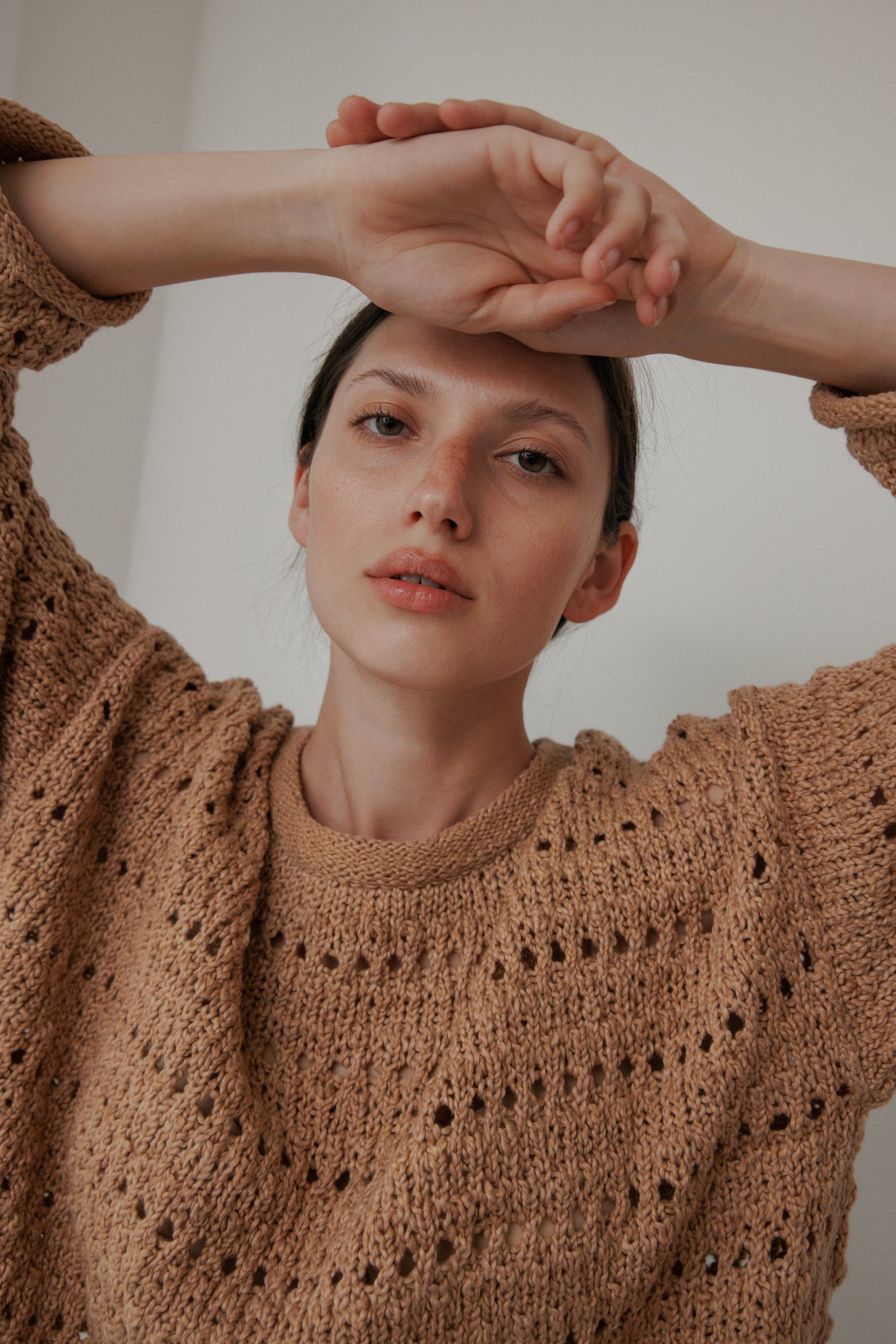 A person wearing the Wol Hide Open Pullover in Tannin poses with arms above their head. They have a neutral expression and short dark hair, standing against a plain light-colored background.