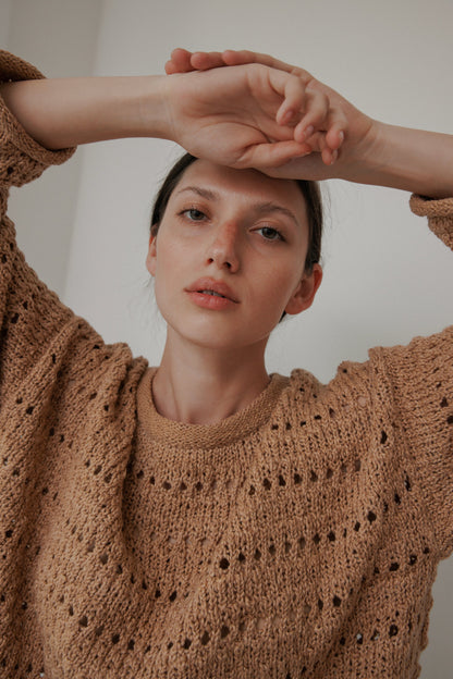 A person wearing the Wol Hide Open Pullover in Tannin poses with arms above their head. They have a neutral expression and short dark hair, standing against a plain light-colored background.