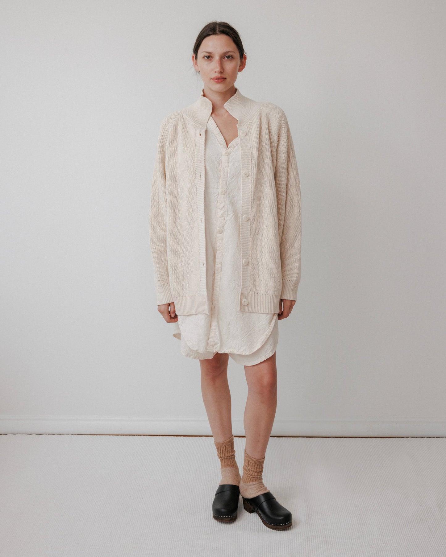 A person stands against a plain white wall wearing Wol Hide's Collar Cardigan in Natural over a light dress, with tan socks and black slip-on shoes. Their hair is pulled back, displaying a neutral expression. Crafted from organic cotton, this USA-made cardigan showcases excellent quality.