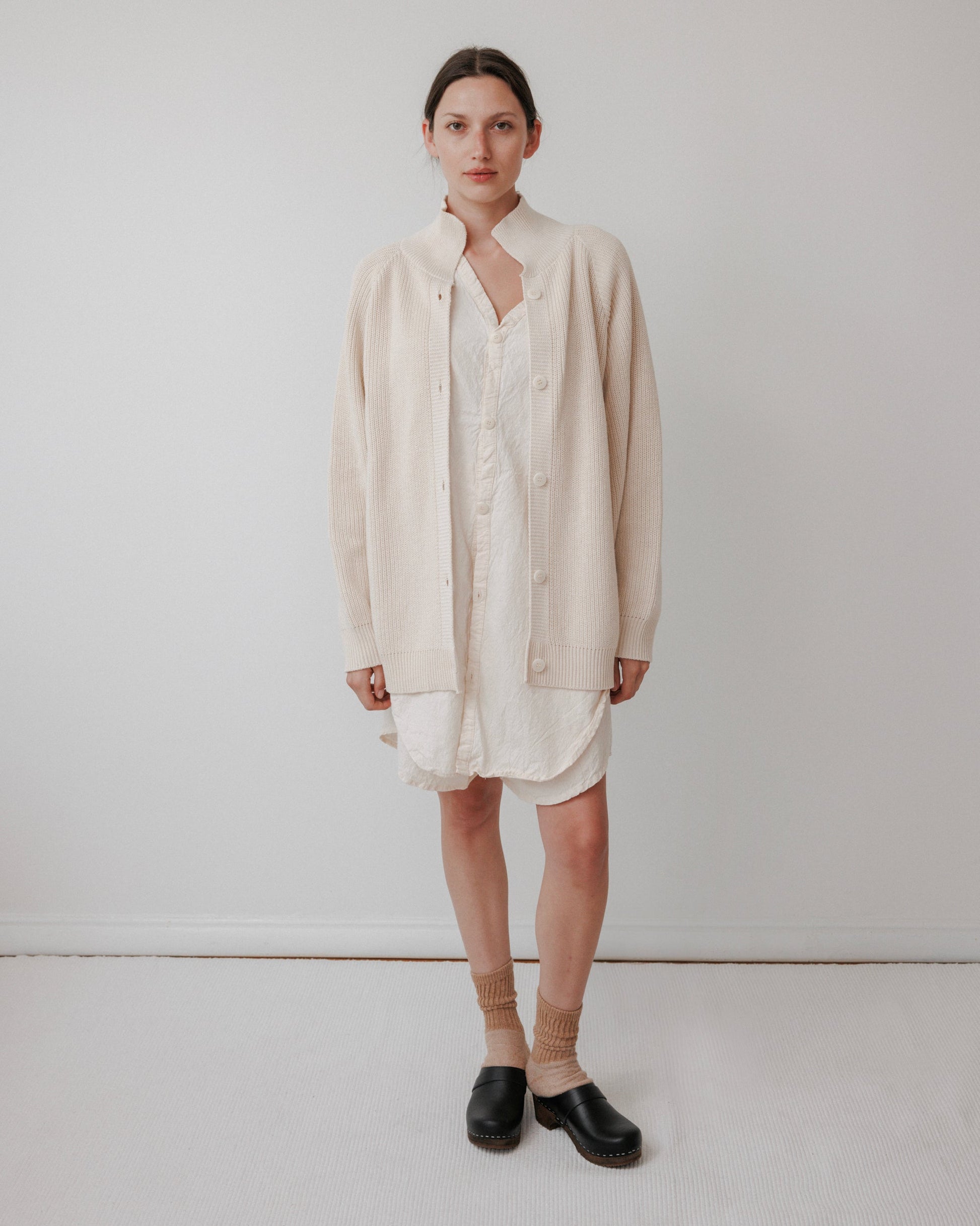 A person stands against a plain white wall wearing Wol Hide's Collar Cardigan in Natural over a light dress, with tan socks and black slip-on shoes. Their hair is pulled back, displaying a neutral expression. Crafted from organic cotton, this USA-made cardigan showcases excellent quality.
