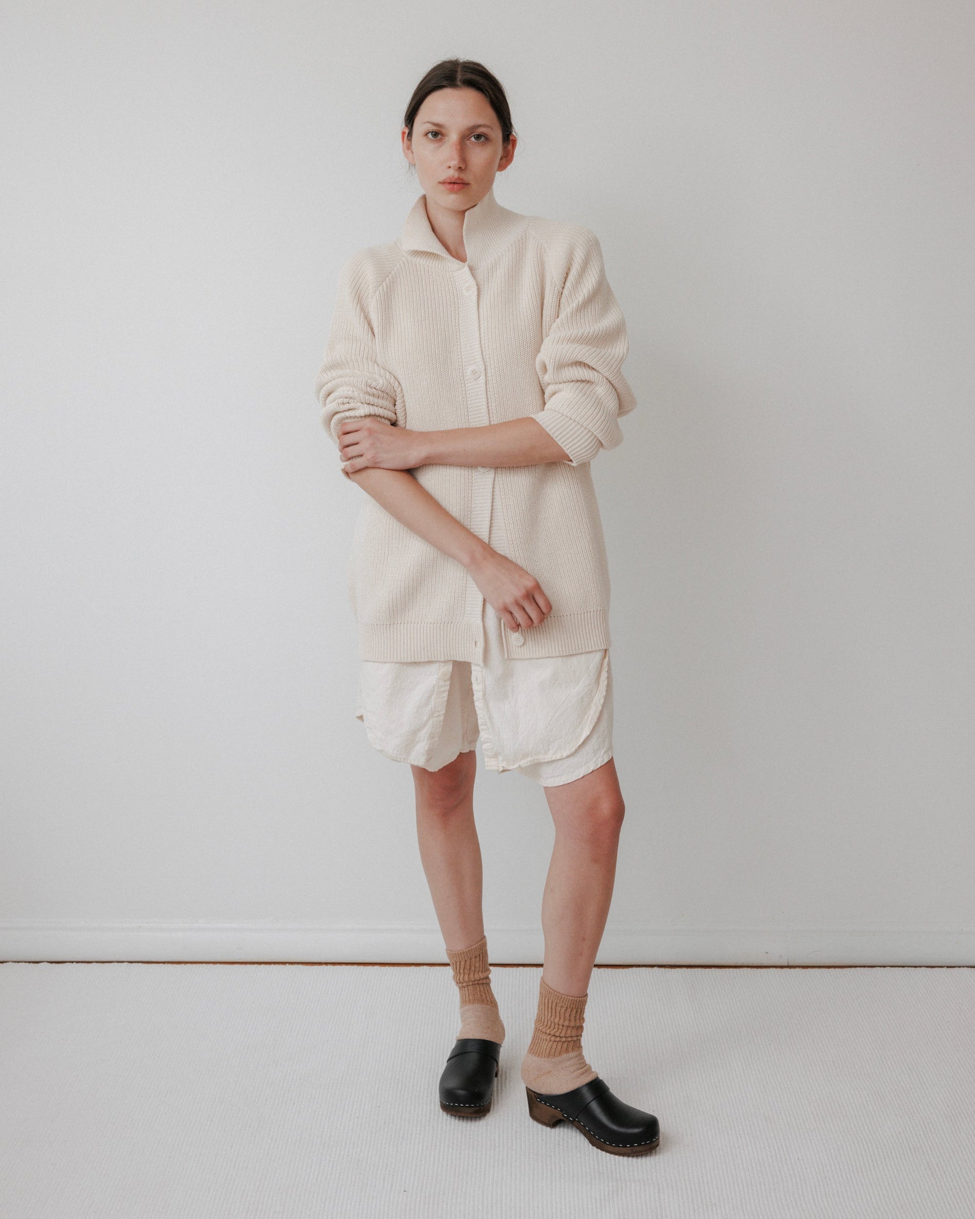A person wearing a long cream Wol Hide Collar Cardigan in Natural over a white dress stands against a plain background. They pair it with beige socks and black clogs, arms lightly crossed and maintaining a neutral expression.
